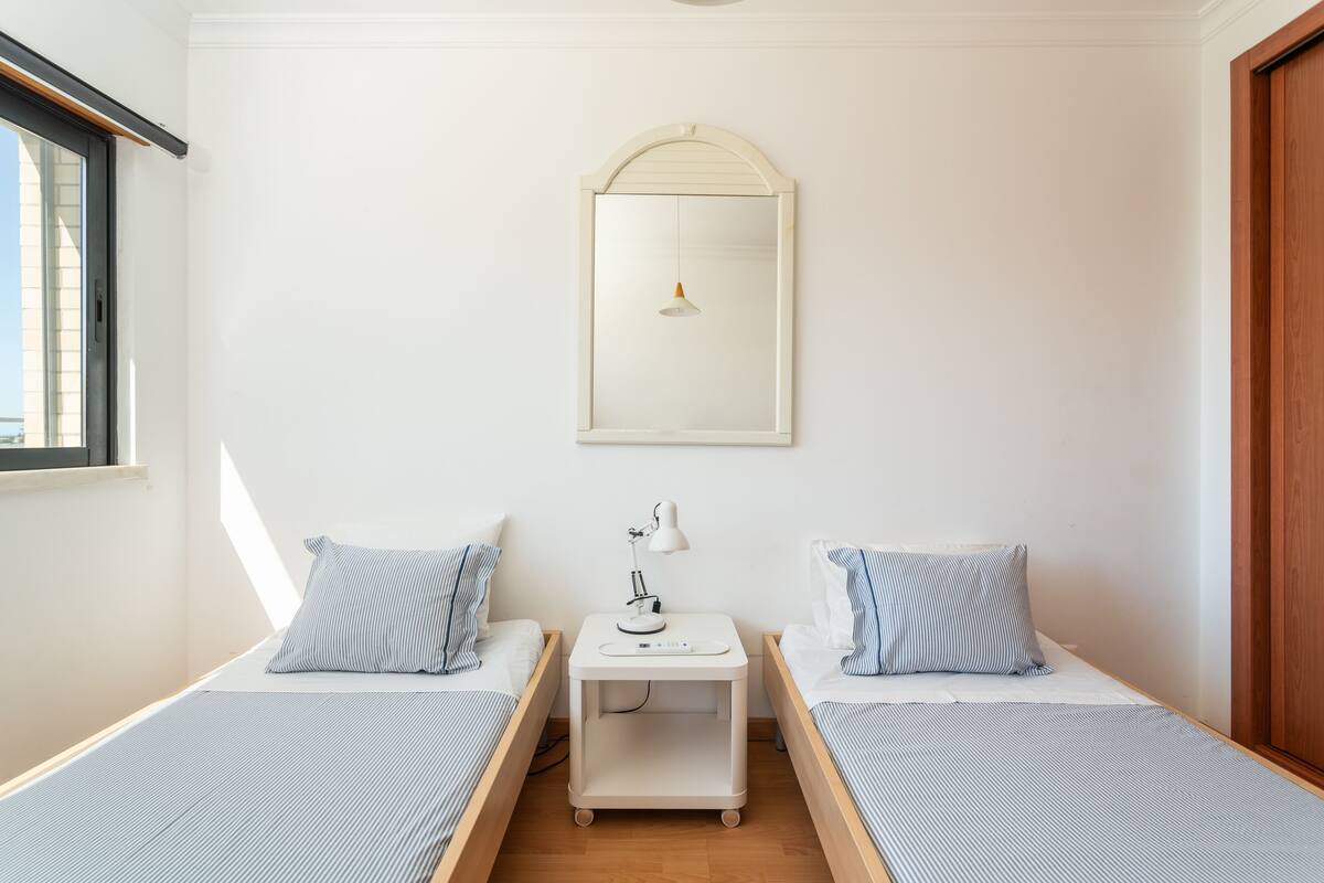 Une chambre à deux lits simples, avec deux lits individuels, une table de chevet avec une lampe, un miroir au-dessus du lit et une fenêtre sur le côté gauche.
