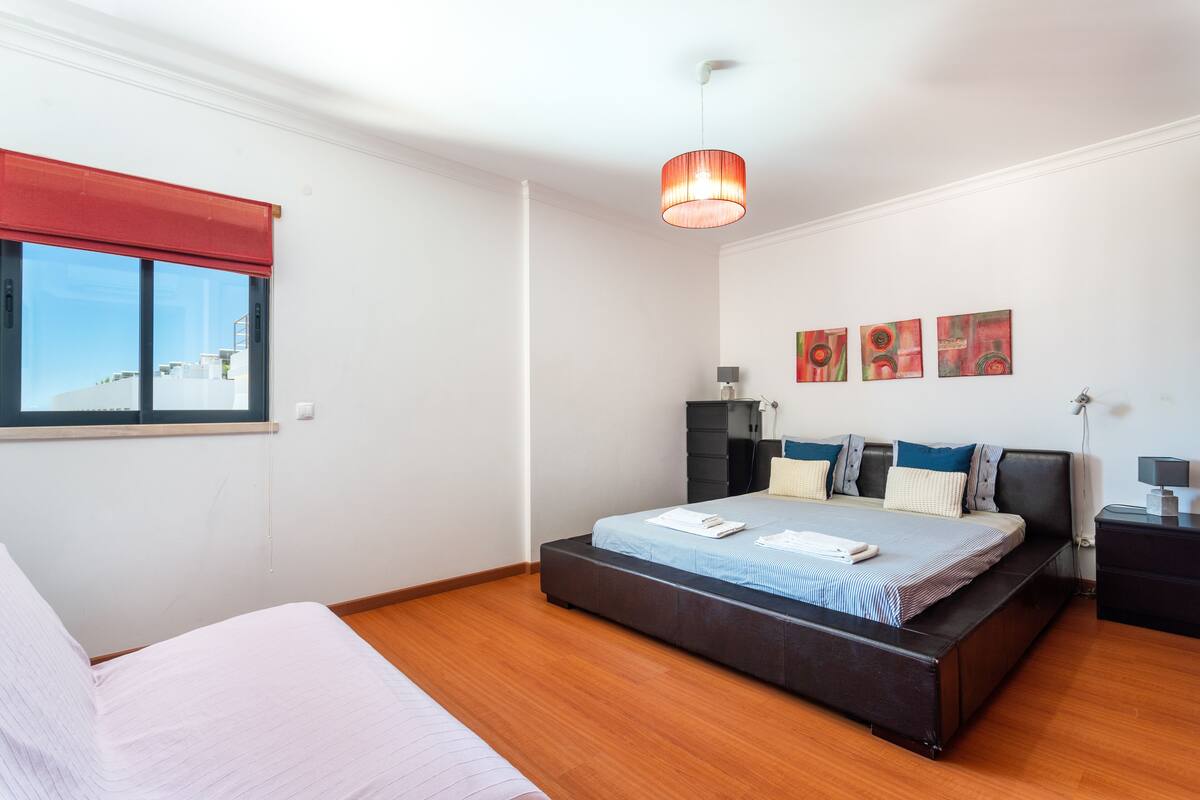 Une chambre moderne avec un grand lit, une fenêtre avec un store rouge et trois tableaux abstraits accrochés au mur.