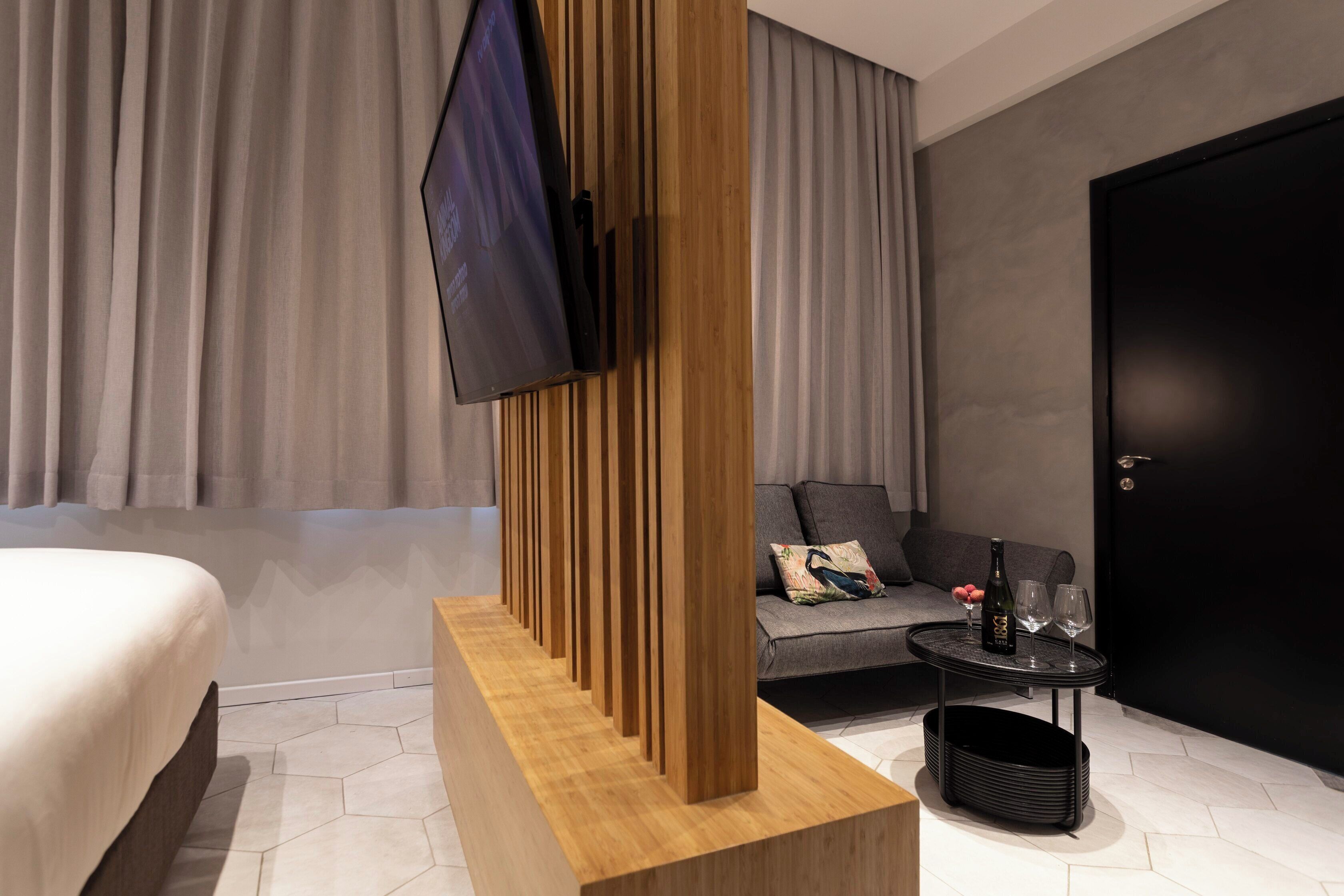 A modern hotel room with a bed, a TV mounted on a wooden panel, a sofa with cushions, a small table with glasses and a bottle, and a door.