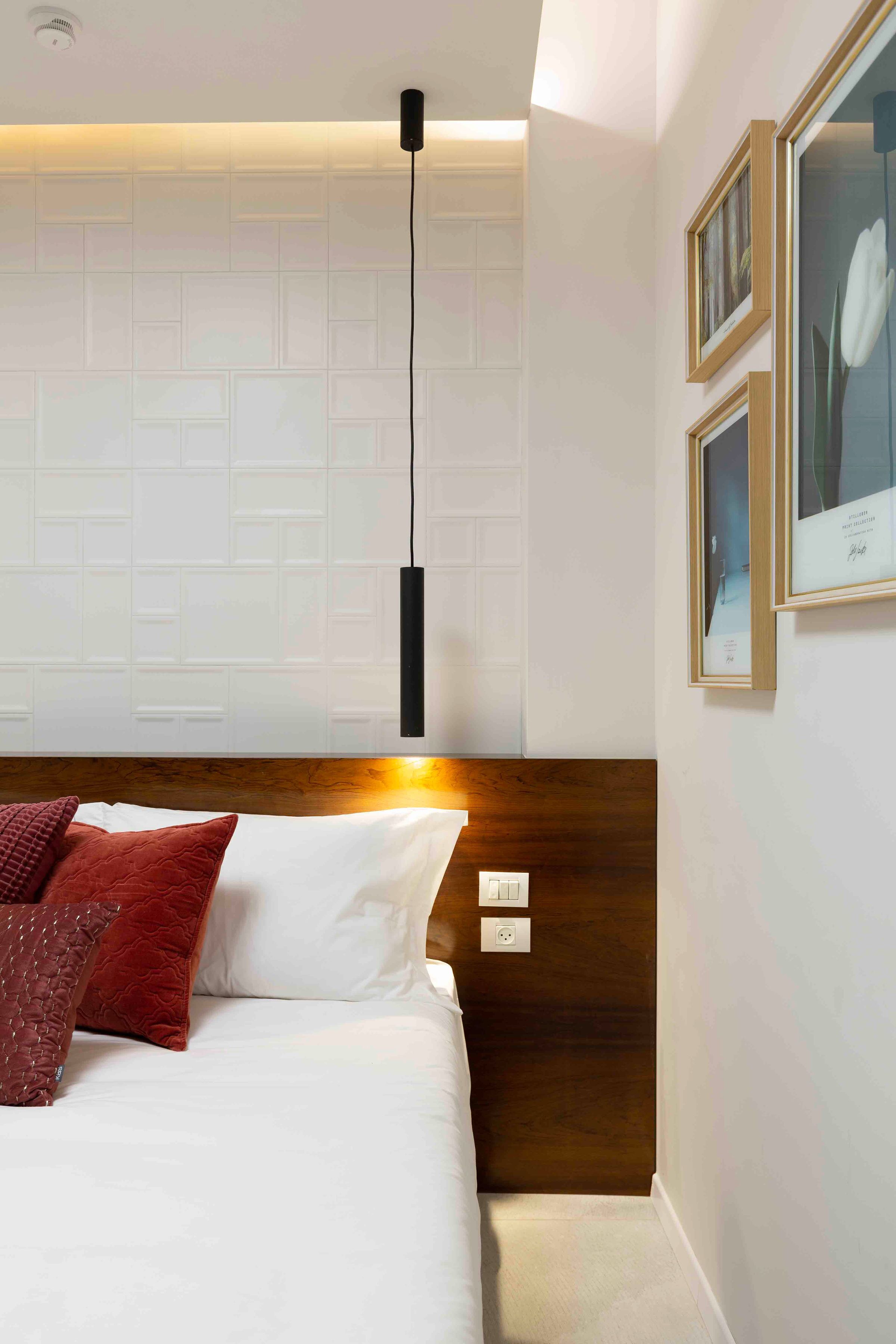 Habitación de hotel con cabecera de madera, cama blanca con almohadas rojas y marrones, lámpara de pared y cuadros en la pared.