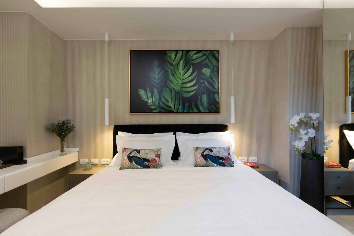 A hotel room with a large bed, two pillows with floral designs, a nightstand with a vase of flowers, and a wall-mounted painting of green leaves.