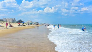 Uma praia com pessoas caminhando e sentadas, edifícios ao fundo e um céu claro.