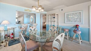 Uma área de refeições com mesa de vidro cercada por cadeiras de vime, um lustre e uma parede azul decorativa.