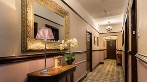 A hallway with a large ornate mirror, a table with a lamp and flowers, and a plaid carpet.