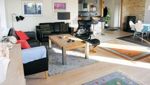 A modern living room with a black leather sofa, a wooden coffee table, and a black leather chair. There is a kitchen area visible in the background with white cabinets and a clock on the wall.