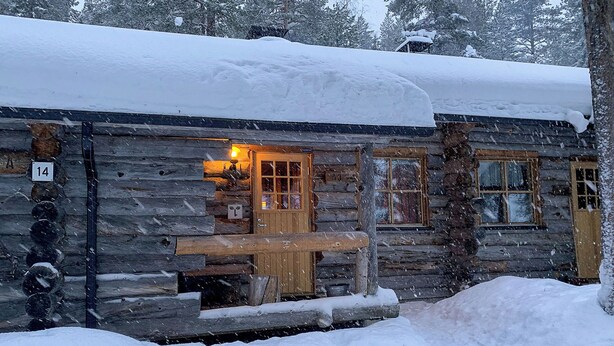 Une cabane en rondins avec un banc en bois à l’extérieur, une fenêtre éclairée et une porte. La neige tombe et la cabane est entourée d’arbres.