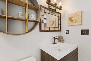 Ein Badezimmer mit einem weißen Waschbecken, einem Spiegel mit einem Hahn-Gemälde und einer Holzwand mit Gläsern.