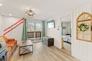 Ein Zimmer mit Holzboden, einer Couch, einem Tisch, einem Koffer, einem Fernseher und einer Tür zu einem anderen Raum.