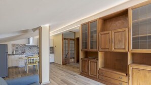 Ampla sala de estar com um grande armário de madeira, um sofá cinzento e uma cozinha ao fundo.