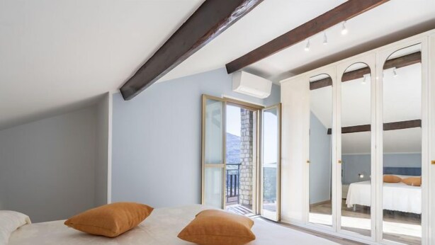 Quarto com uma cama grande, duas almofadas, uma viga de madeira no teto e uma porta de vidro deslizante para outro cômodo.