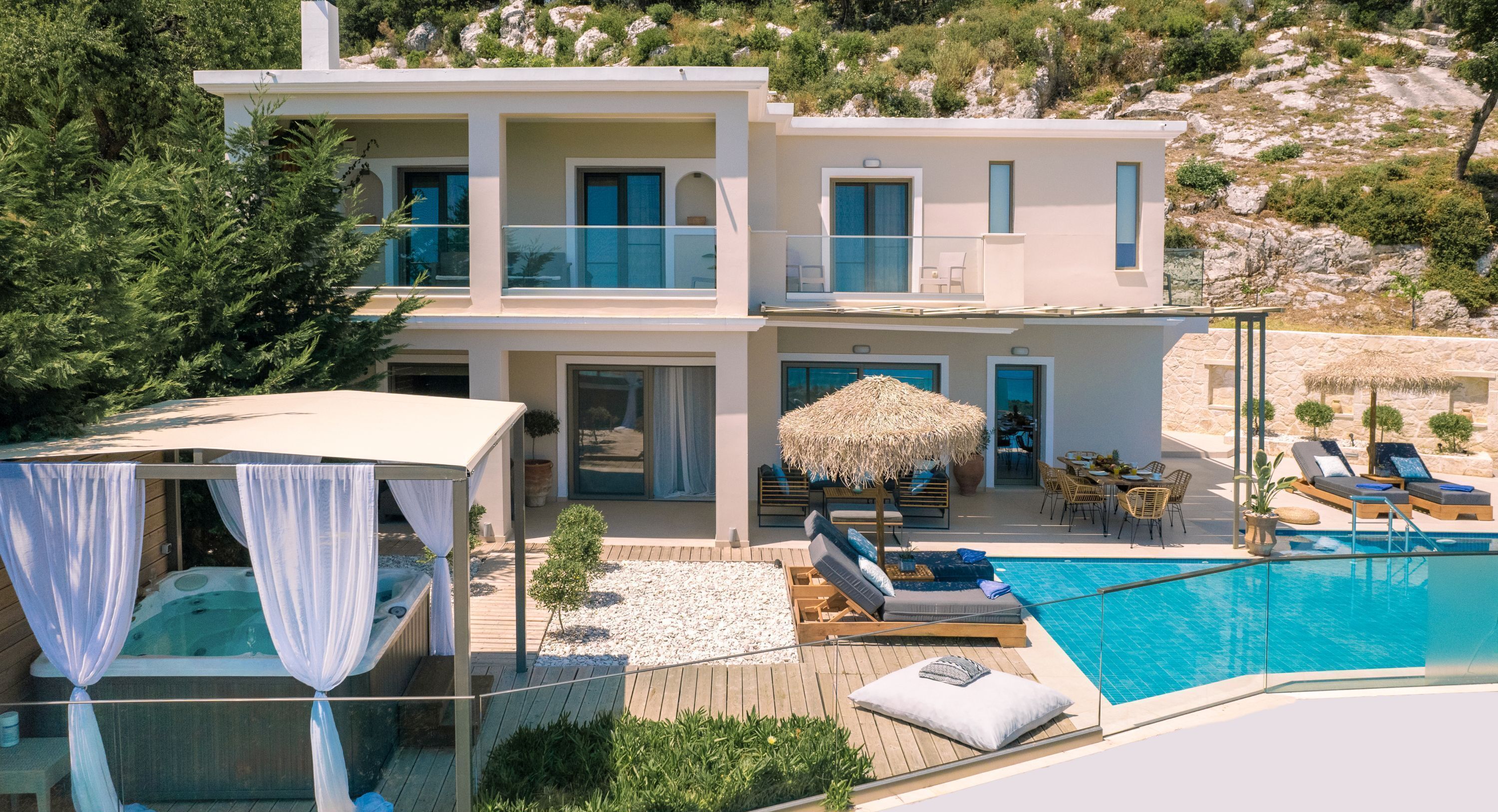 En moderne villa i to etager med swimmingpool, udendørs møbler og en stråtækt parasol.