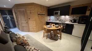 A cozy living room with a sofa and a small Christmas tree, a wooden dining table with chairs, and a kitchen area with modern appliances.