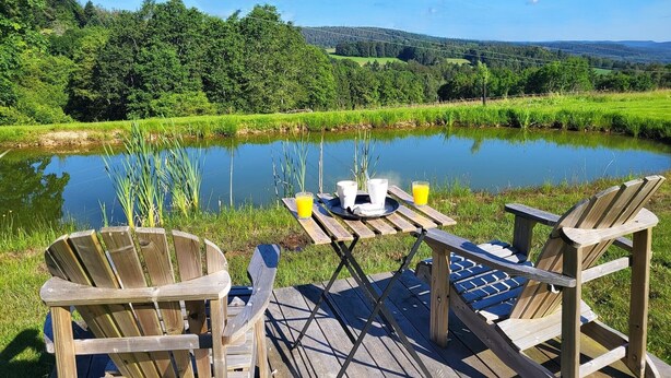 A wooden deck with two Adirondack chairs overlooking a pond surrounded by lush greenery and hills. - Nature Cottage Vosges Le Renard (Anould)