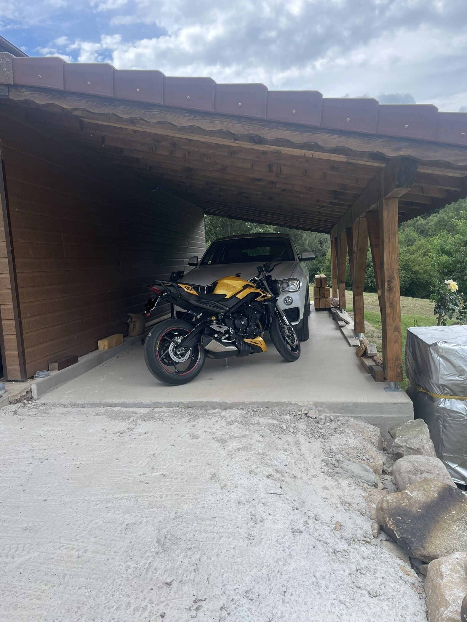 A covered parking area with a motorcycle and a car parked under a wooden structure.