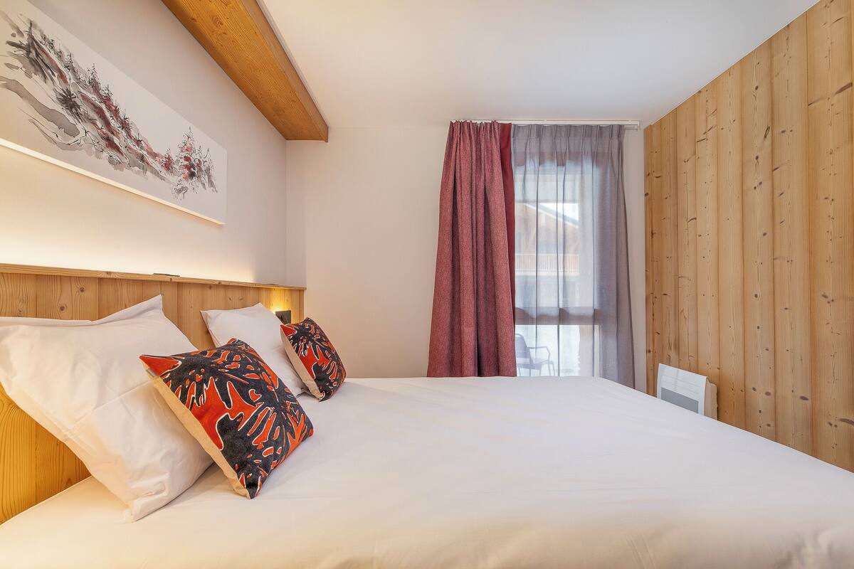 Une chambre à coucher avec une tête de lit en bois, un grand lit avec des oreillers blancs et à motifs, une fenêtre avec des rideaux et un panneau mural en bois.