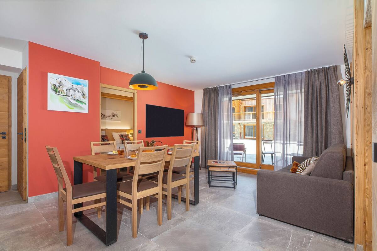 Un salon comprenant un coin repas, un canapé et une table avec des chaises. Il y a un tableau au mur et une fenêtre avec des rideaux.