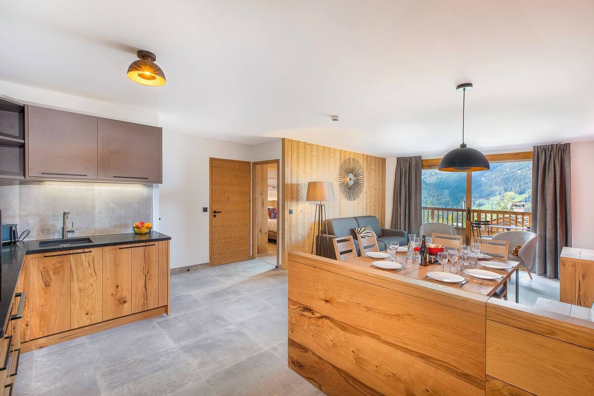 Une cuisine moderne avec des meubles en bois, un coin repas offrant une vue sur les montagnes et un salon chaleureux.