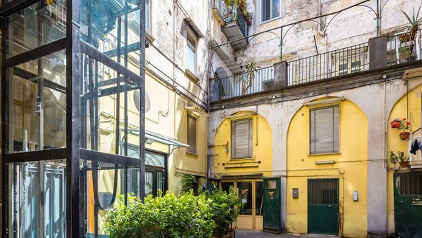 Un patio con un edificio de vidrio, suelo empedrado de piedra y edificios amarillos con puertas verdes.