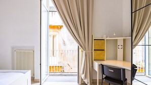Un dormitorio moderno con cama, escritorio y silla. Hay una ventana con cortinas, un radiador y vistas al exterior.