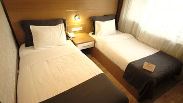 Quarto de hotel com duas camas de solteiro, cabeceira de madeira, mesas de cabeceira com luminárias e uma janela com cortinas leves.