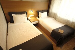 A hotel room with two single beds, a wooden headboard, bedside tables with lamps, and a window with sheer curtains.