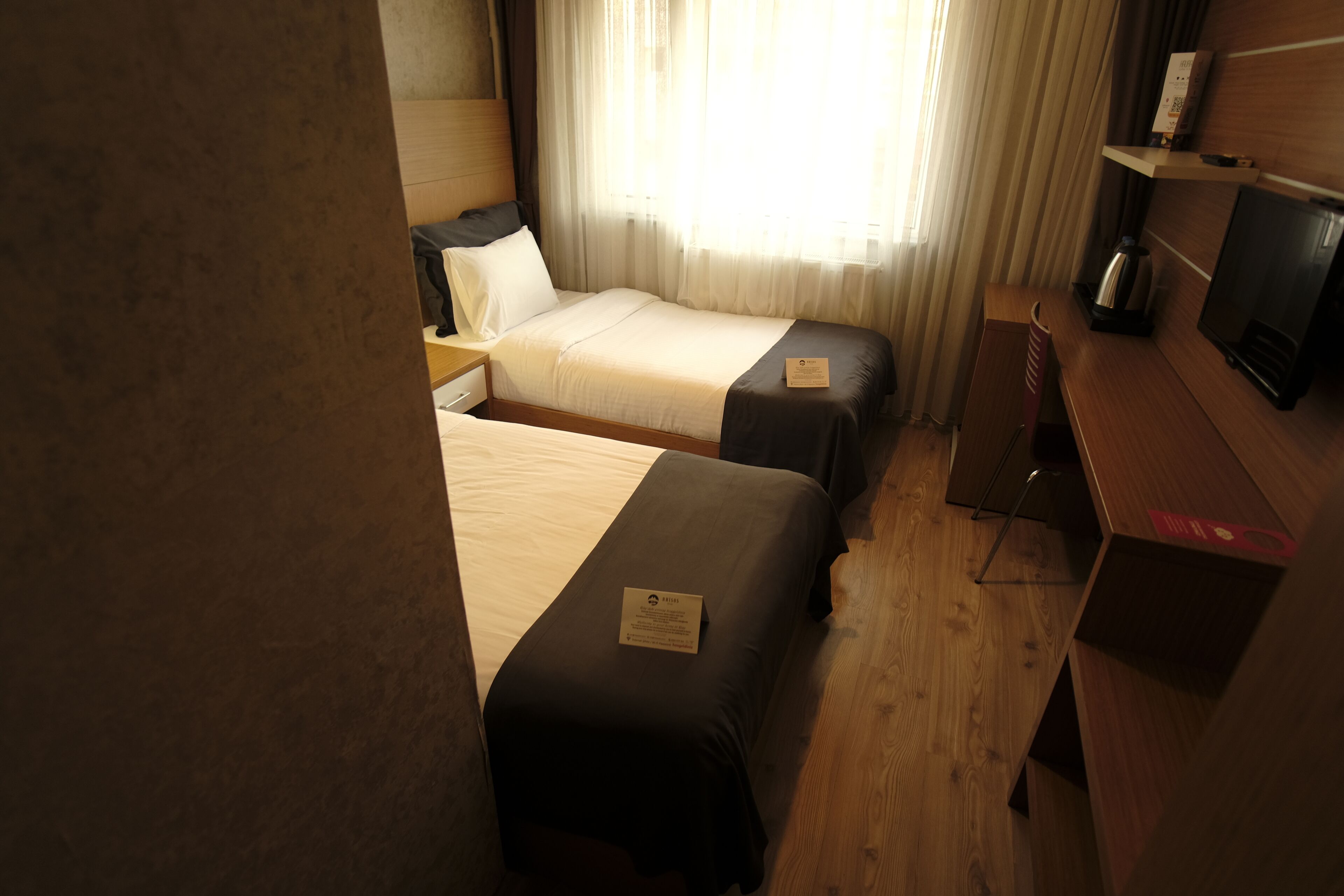 A hotel room with two beds, a wooden desk, a television, and a window with curtains.