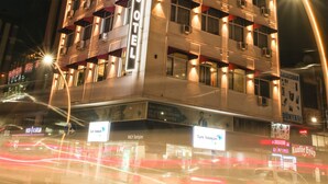 Een meerlaags hotel met verlichte ramen en balkons, een zichtbaar bord met 'Rhisos Hotel' en een straatbeeld met passerend verkeer.