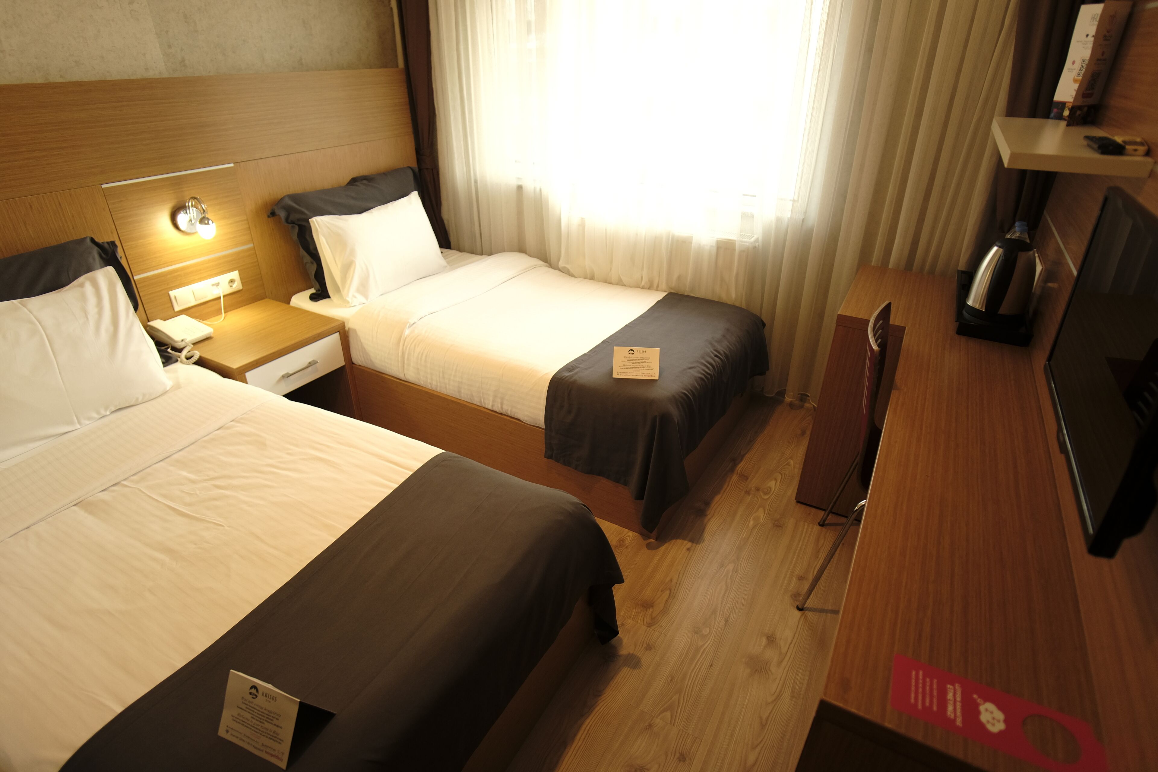 Habitación de hotel con dos camas, una mesita de noche con lámpara, un televisor y un estante con objetos.