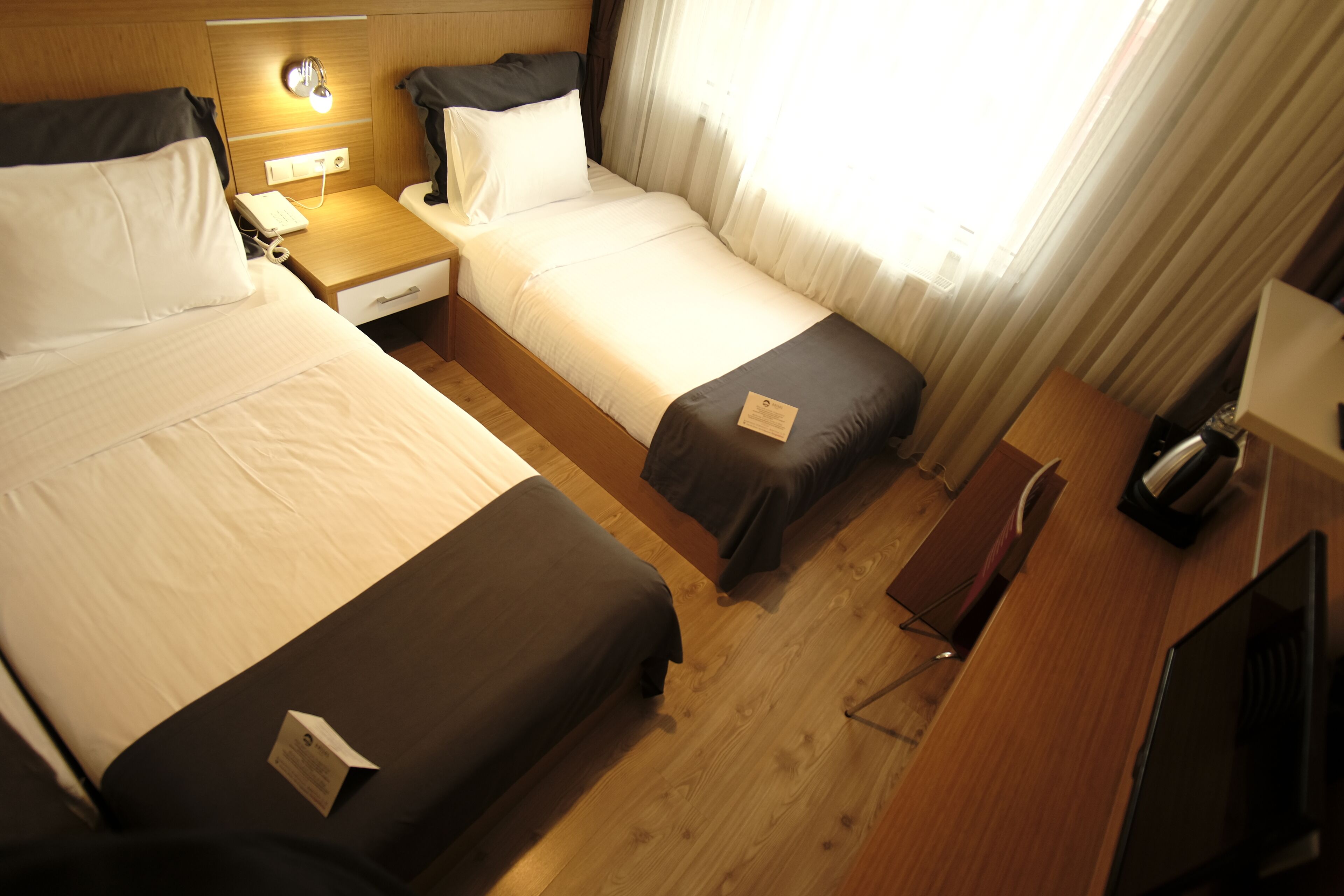 A hotel room with two single beds, a wooden desk, a television, and a nightstand with a lamp.