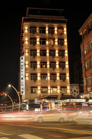 A multi-story hotel with illuminated windows and balconies, a visible sign reading "RHSOS HOTEL," and a street with passing vehicles.