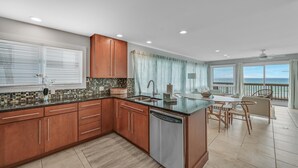 A modern kitchen with wooden cabinets, a dishwasher, and a dining area with a view of the ocean. - Gulf-Front with Private Deck and Water Views (Miramar Beach)