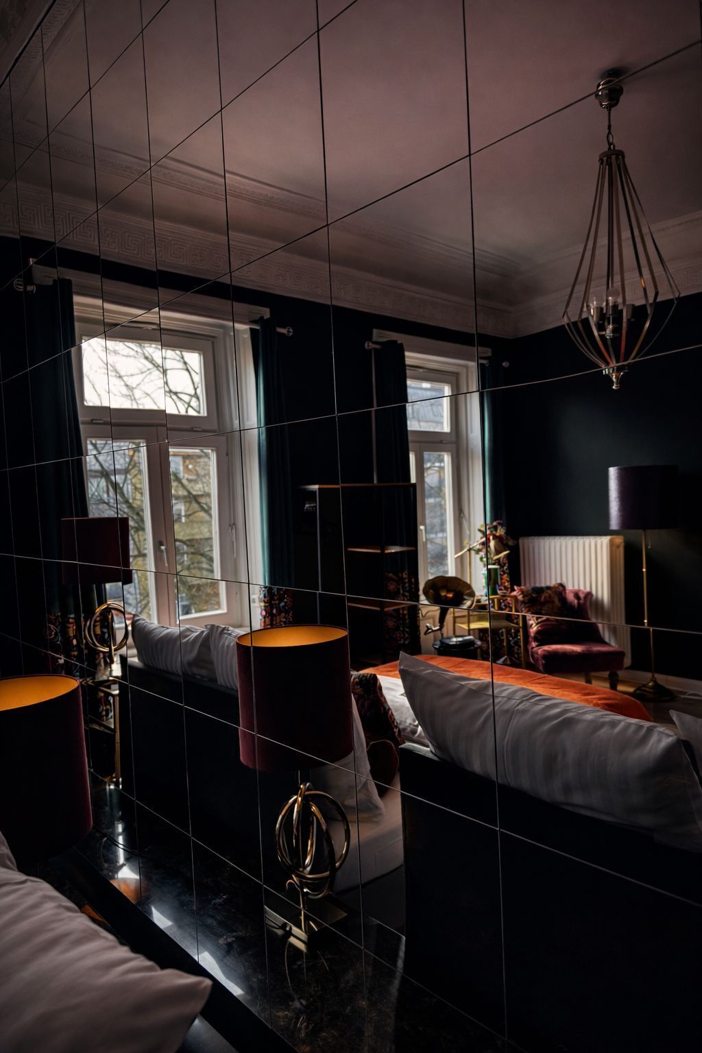 A modern bedroom with a large bed, a desk, and a chair. There are windows with curtains, a chandelier, and a floor lamp.