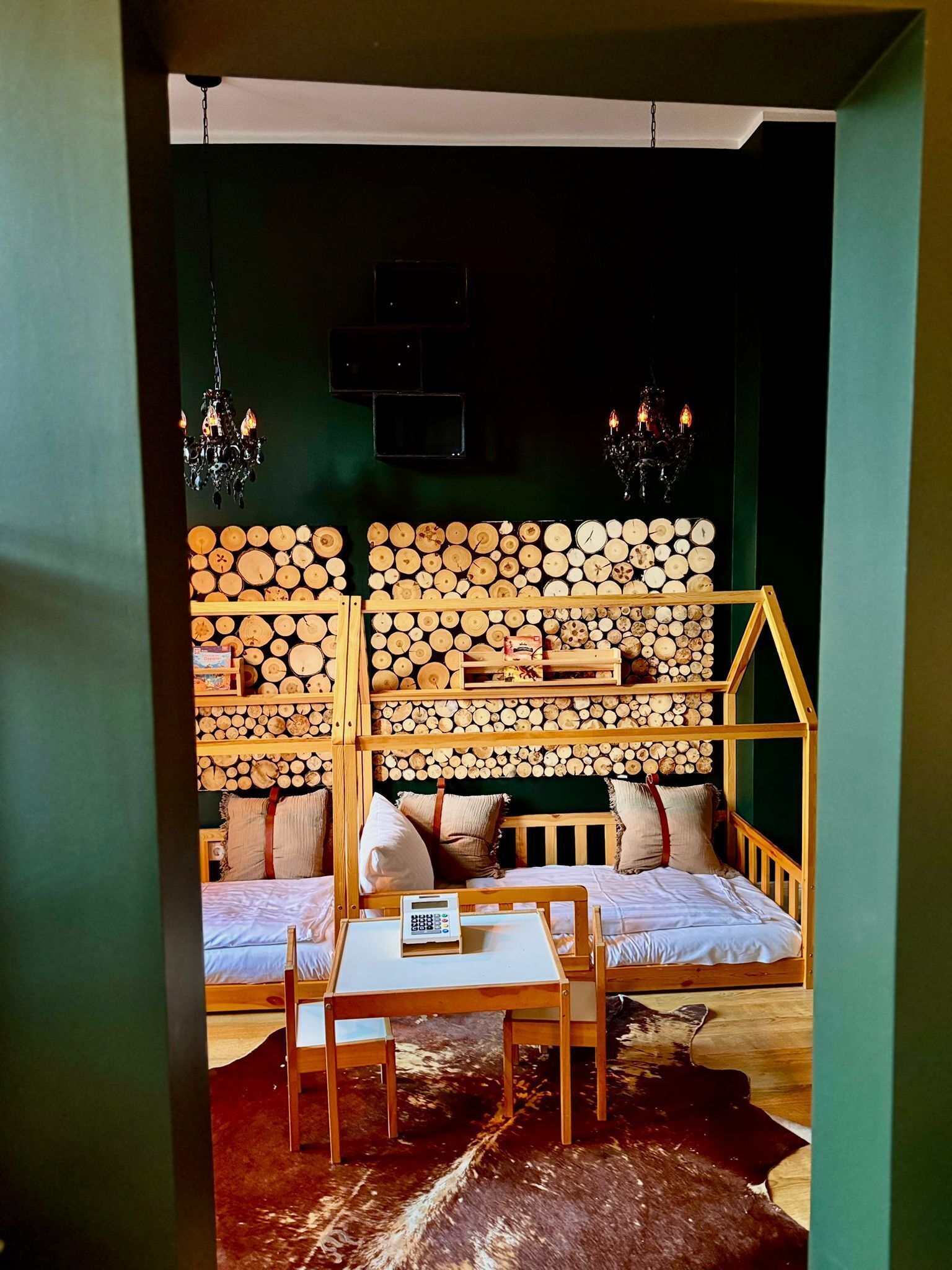 A cozy room with a bed, a small table, and a wooden bench. There is a wall decorated with stacked logs and two chandeliers.