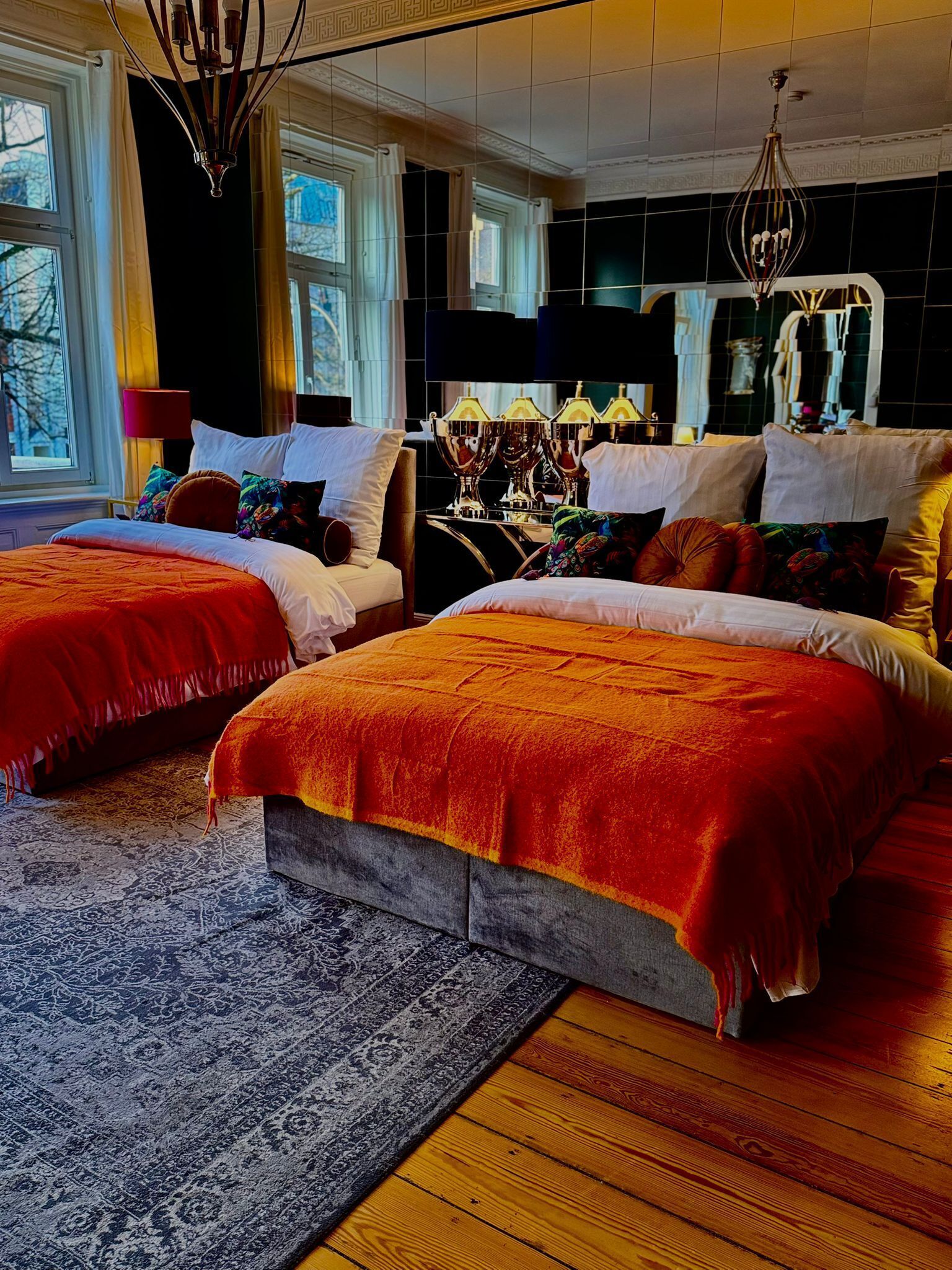 A hotel room with two beds, a large window, a chandelier, and a mirror.
