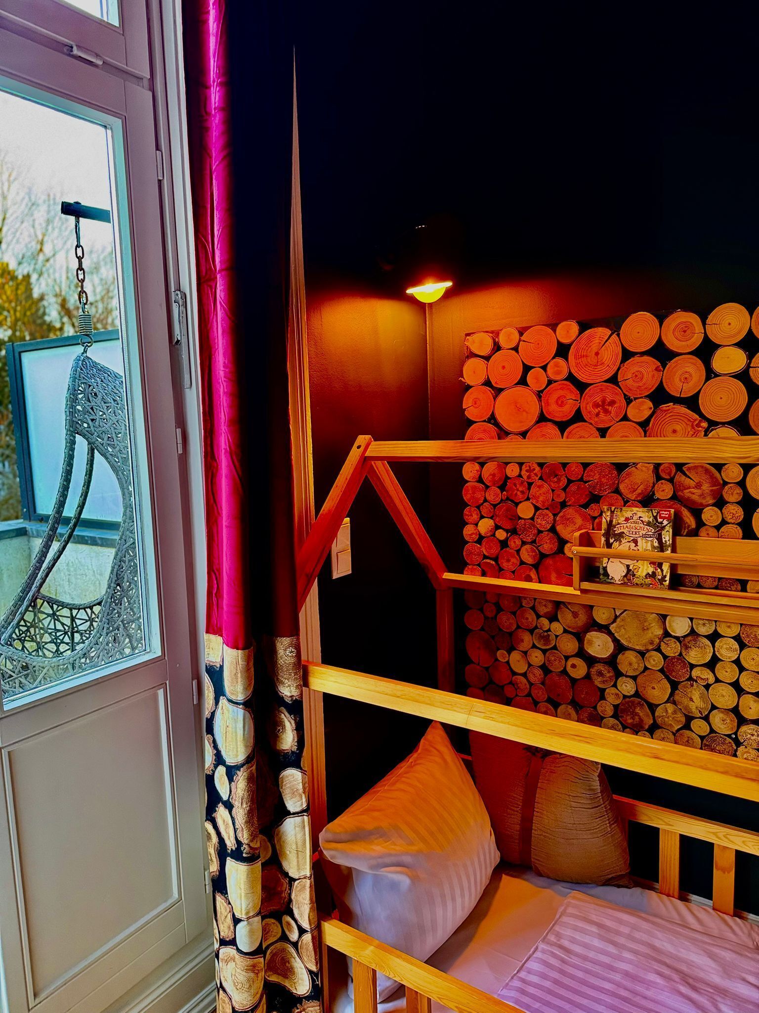 A cozy room with a wooden bunk bed, a window with a decorative curtain, and a wall adorned with stacked logs.