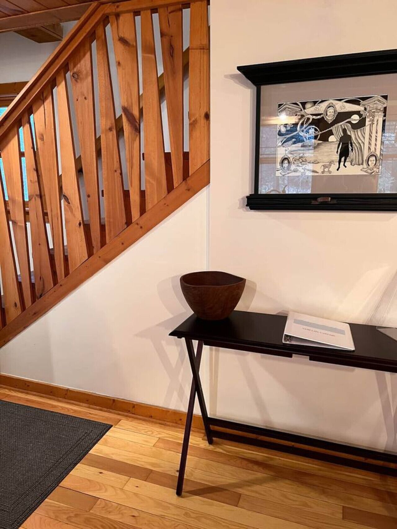 A wooden staircase with a black railing, a framed artwork on the wall, a small table with a bowl, and a mat on the floor.