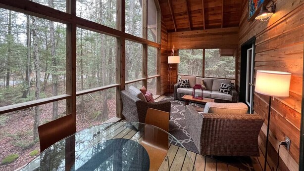 Interior acolhedor de uma cabana com uma mesa de vidro, móveis de vime e uma grande janela com vista para a floresta.