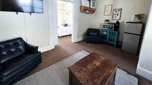 Un espacio habitable compacto con un sofá de cuero negro, una mesa de centro de madera, una kitchenette con microondas y refrigerador, y una puerta que da a un dormitorio.