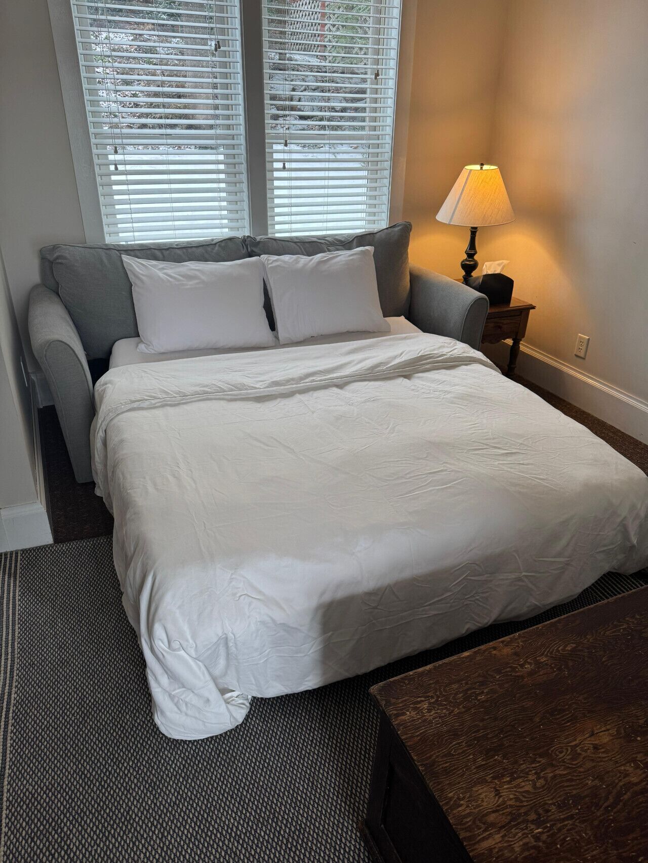 Een net opgemaakt bed met witte lakens, een grijze bank, een klein houten nachtkastje met een lamp, en een raam met jaloezieën.