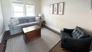 Una sala de estar con un sofá gris, una mesa de centro de madera, un sillón negro y una alfombra estampada.