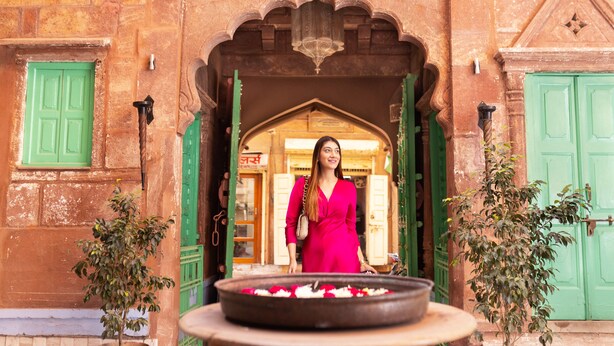 A woman in a pink dress standing in front of a traditional building entrance with green doors and a decorative archway.