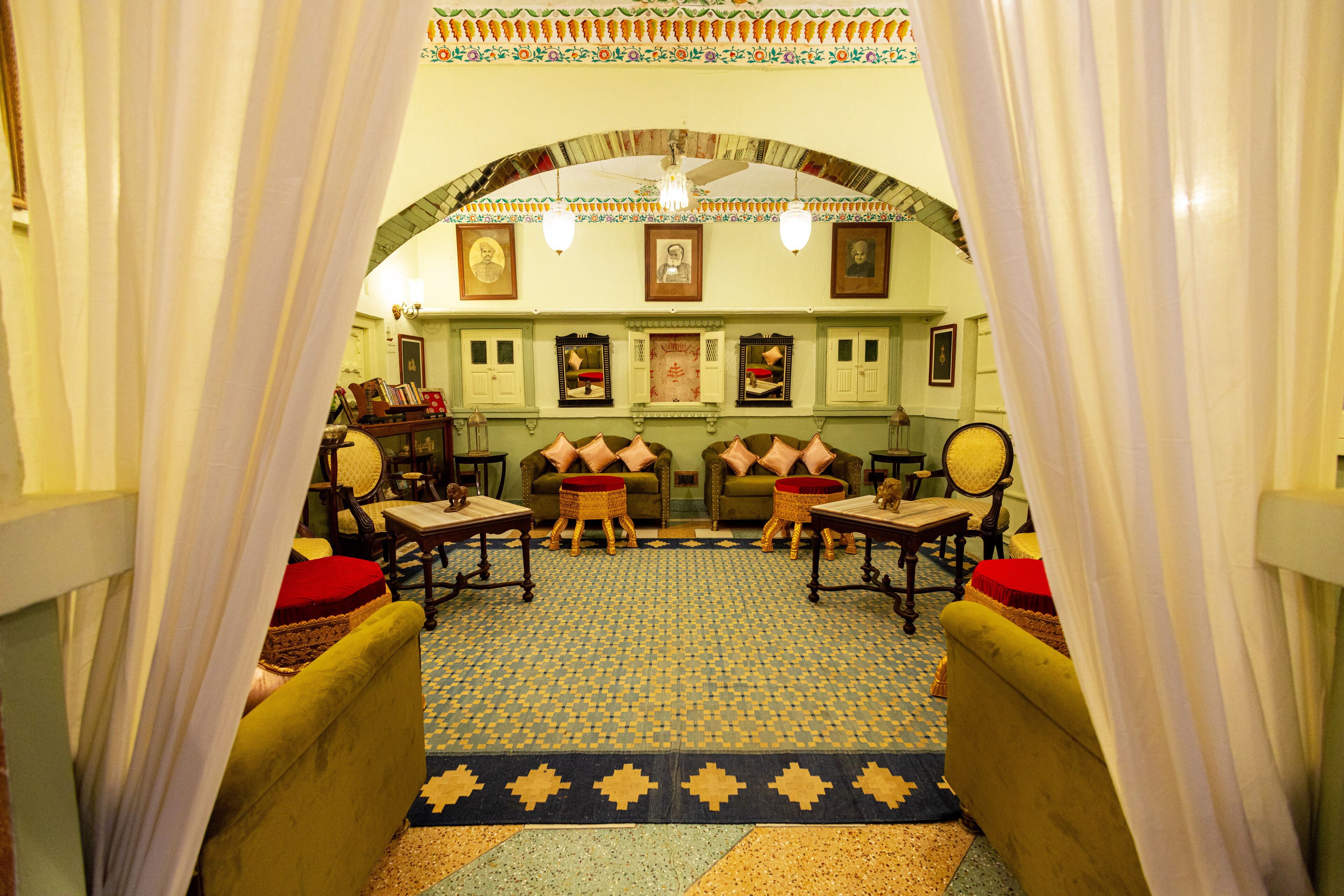 A room with a patterned floor, green sofas with red cushions, wooden chairs, and a ceiling with a decorative arch and framed pictures.