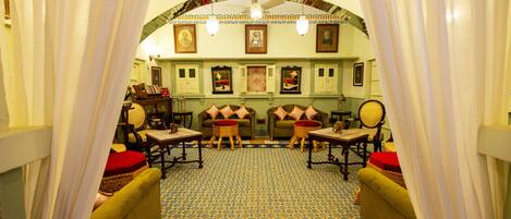 A room with a patterned floor, green sofas with red cushions, wooden chairs, and a ceiling with a decorative arch and framed pictures.