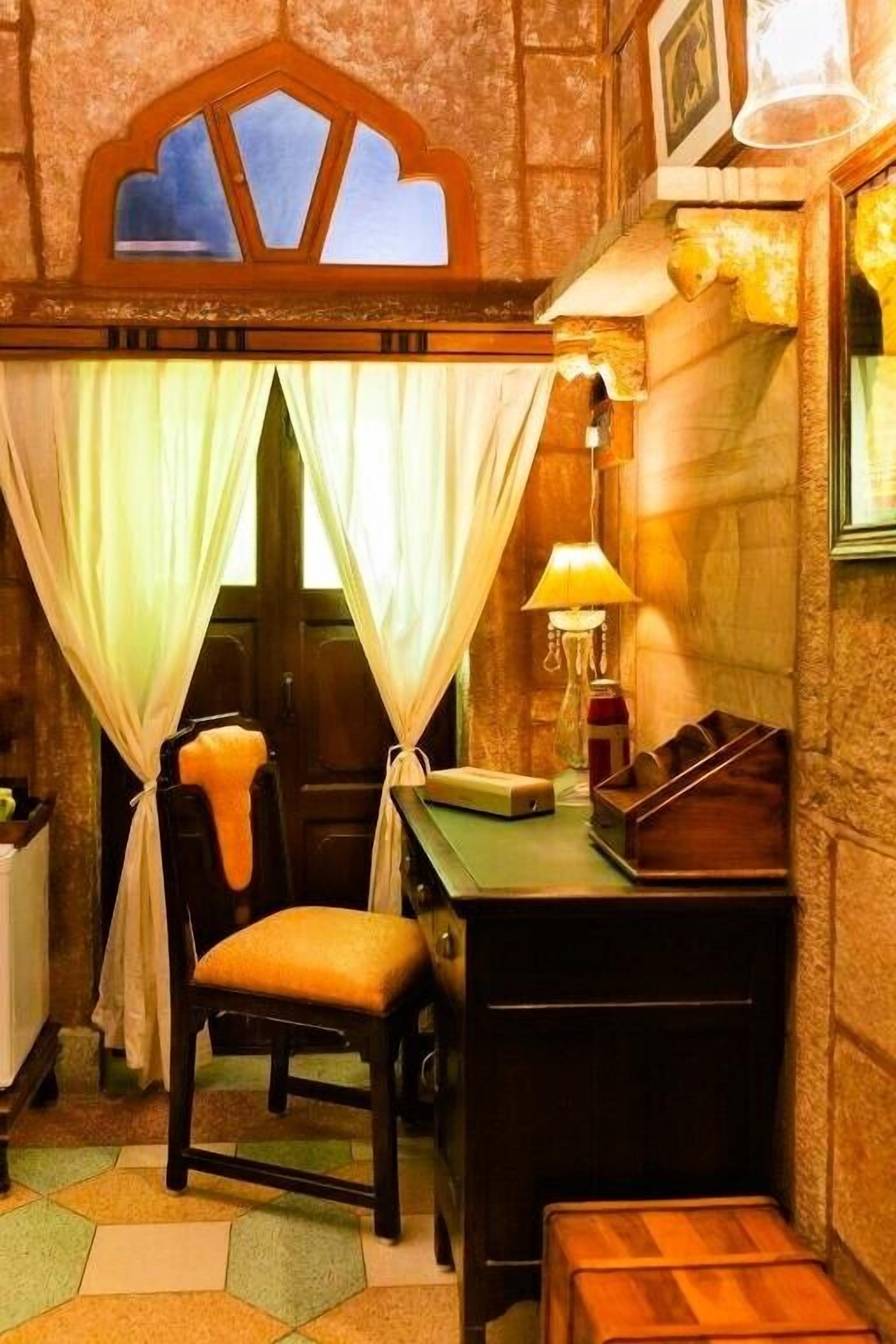 A cozy room with a wooden desk, a chair, a lamp, and a window with curtains.