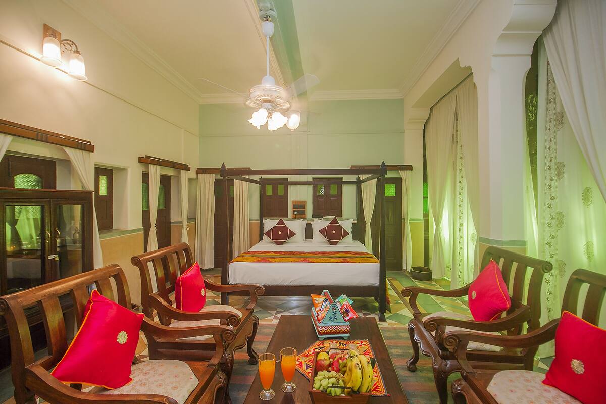 Ein Himmelbett mit weißer Bettdecke und roten Kissen, Holzstühle mit roten Kissen, ein Obstkorb und ein Tisch mit Gläsern Orangensaft.