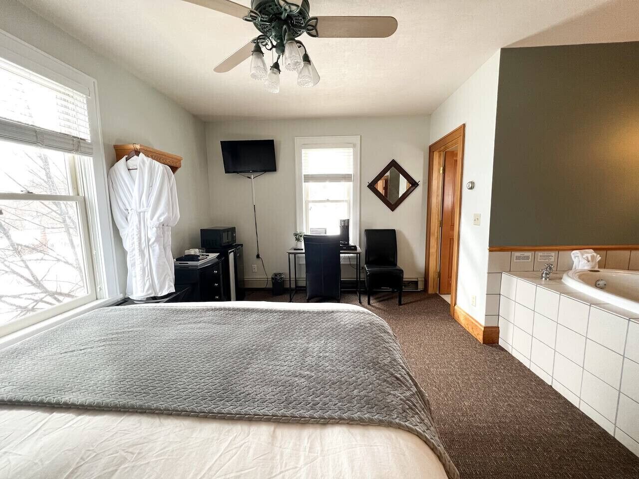 Un dormitorio con una cama grande, un escritorio, una silla, un televisor, un ventilador de techo, una ventana y un baño con bañera.