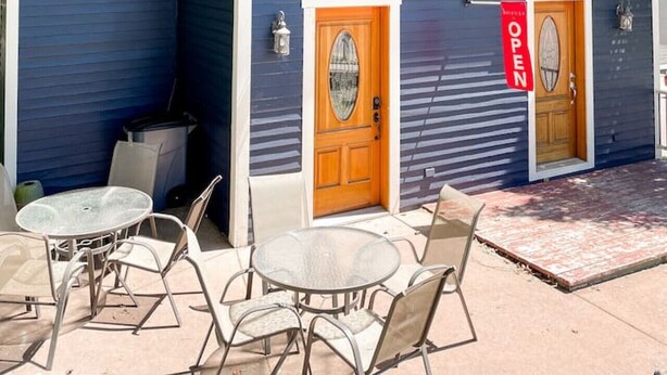 Un bâtiment à deux étages, avec une façade bleue, une porte d’entrée en bois et une petite fenêtre. On y trouve des tables et des chaises à l’extérieur, ainsi qu’une enseigne rouge indiquant « OUVERT ».