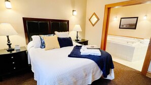 Une chambre d’hôtel avec un grand lit, deux tables de chevet avec des lampes, une table de nuit, une salle de bain avec une baignoire et un tableau encadré au mur.