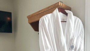 Une robe de chambre blanche est suspendue à un cintre en bois dans une pièce où se trouve une petite table sur laquelle sont posés des articles de toilette et des serviettes pliées.