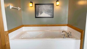 Une salle de bain avec une grande baignoire, deux appliques murales et un tableau représentant une forêt enneigée.